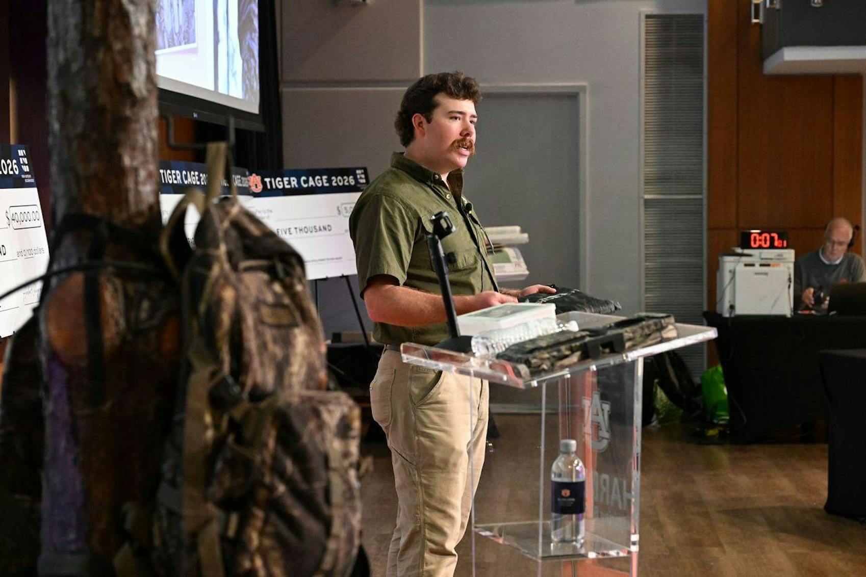 A man in a green shirt stands at a podium presenting, with backpacks and checks visible in the background.