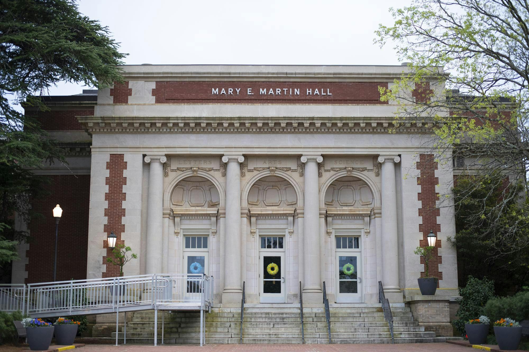A large, classical-style building features three arched doorways with decorative columns, flanked by greenery and colorful flower pots.