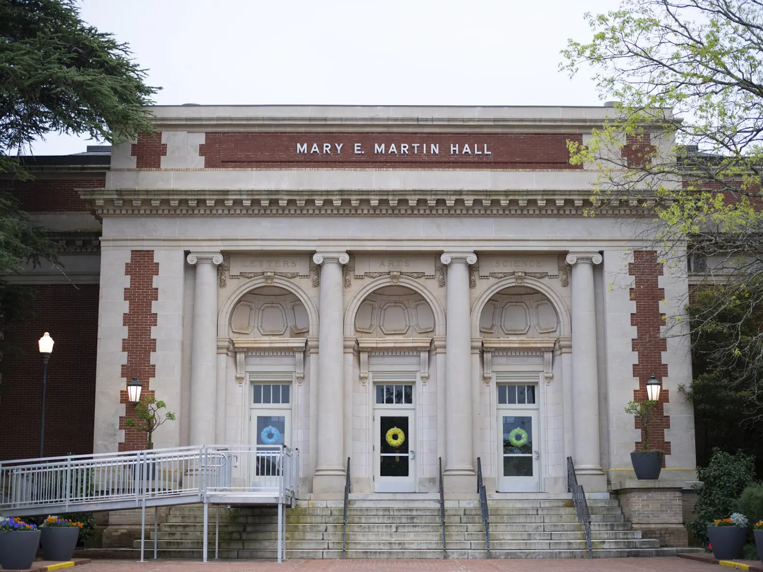 A large, classical-style building features three arched doorways with decorative columns, flanked by greenery and colorful flower pots.