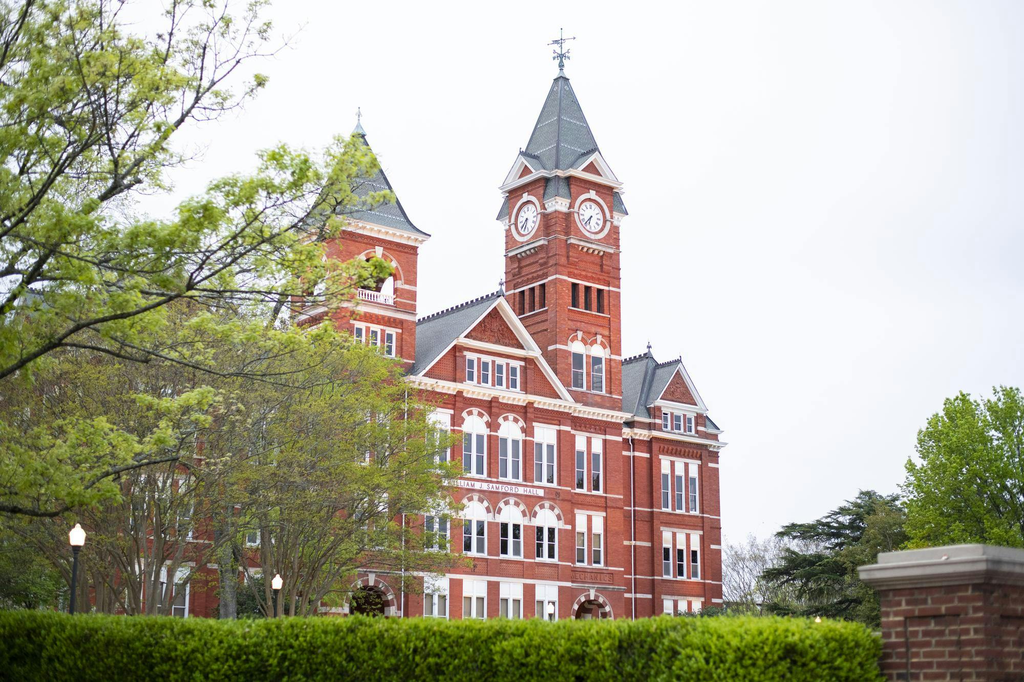 A red brick building features prominent clock towers, surrounded by green trees and neatly trimmed bushes.