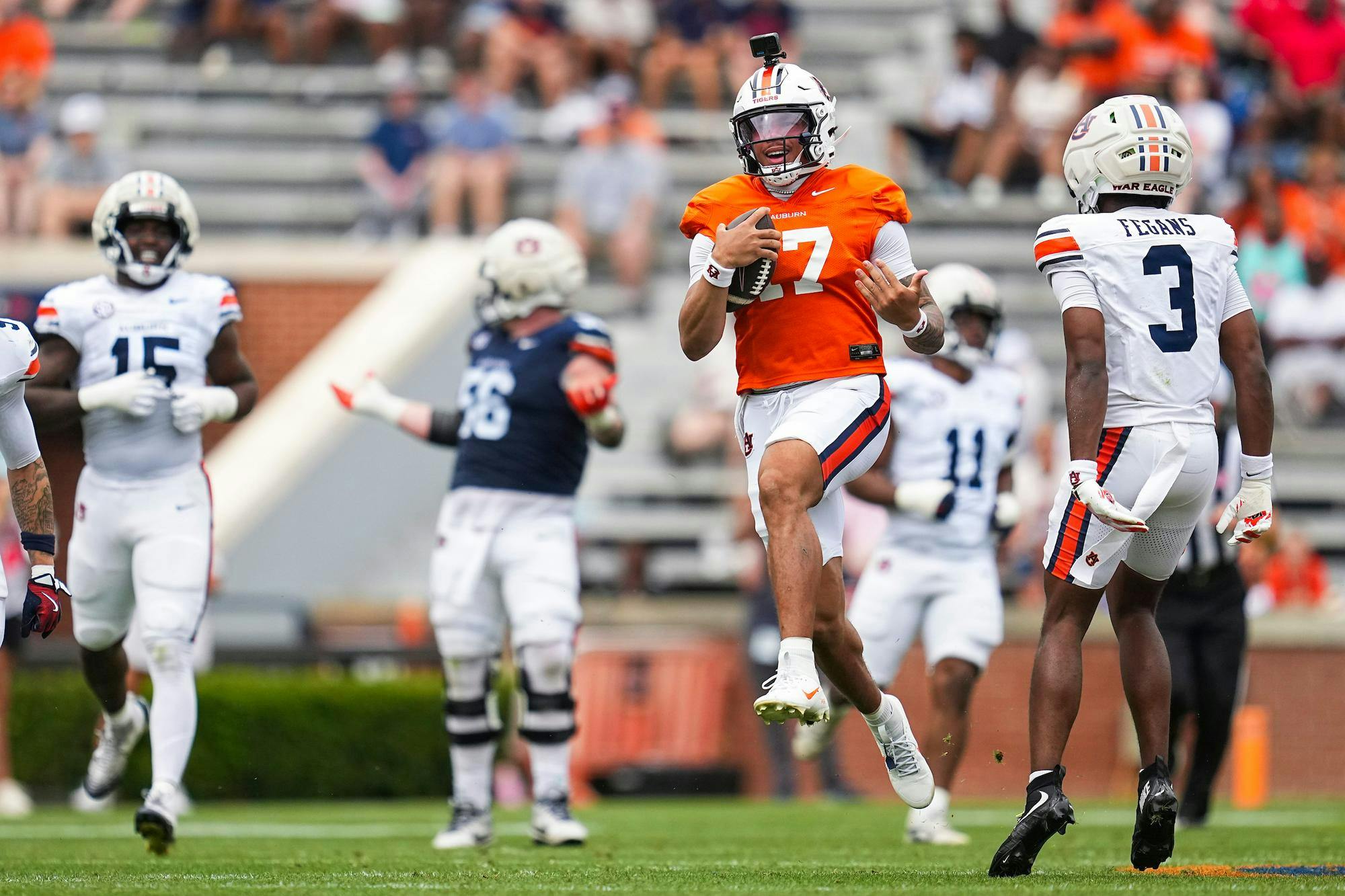 A football player in an orange jersey runs with the ball while two defenders pursue him on a grassy field.