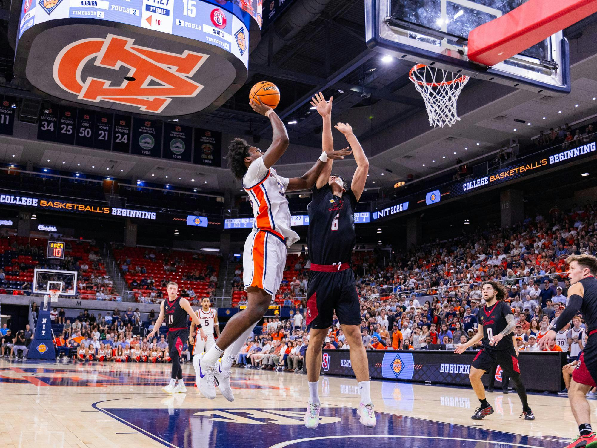 A player in a white and orange jersey leaps to shoot a basketball while defenders in black jerseys attempt to block the shot.