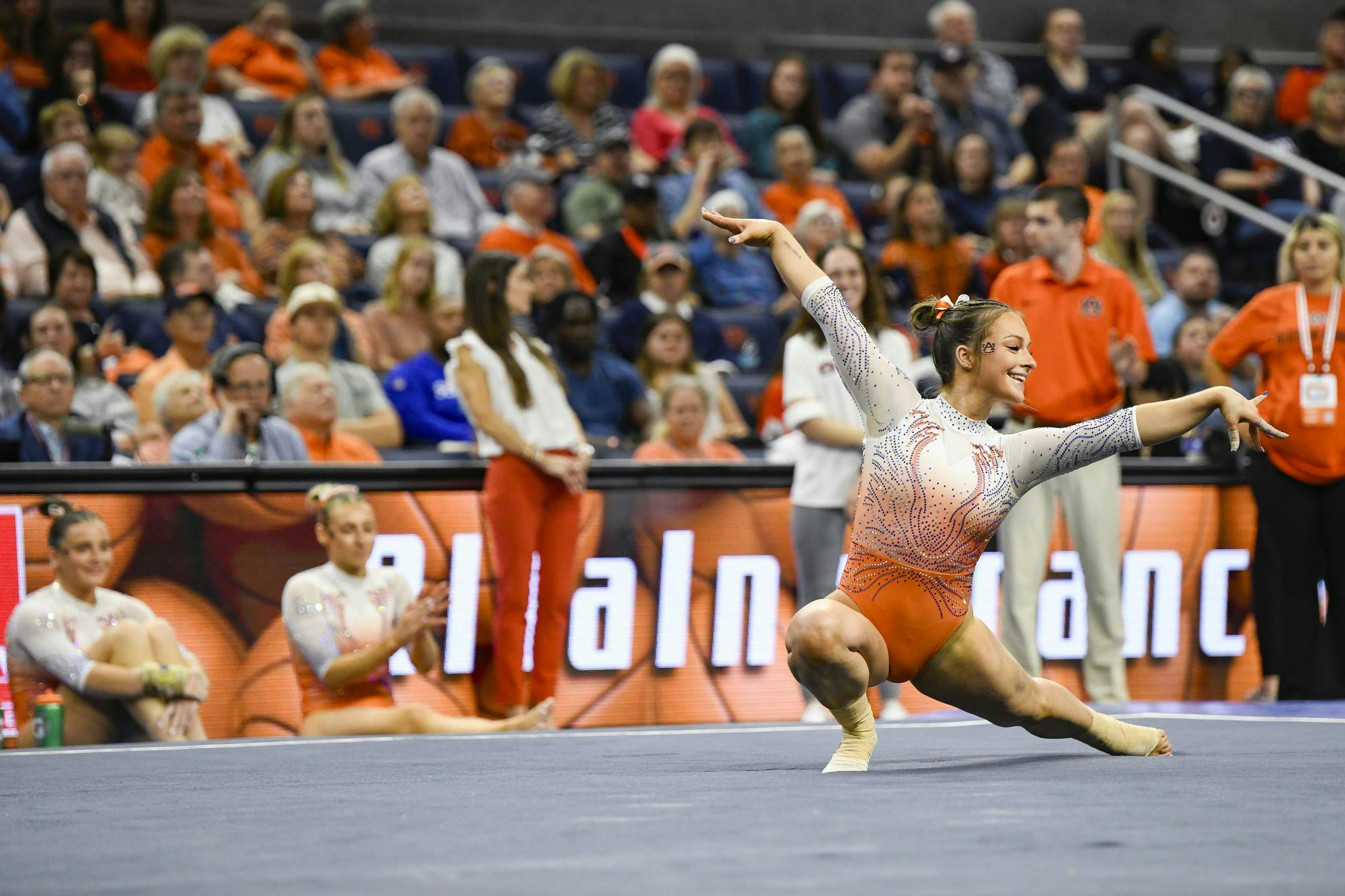 A gymnast in an orange and silver outfit performs a pose while two others sit nearby, with a crowd in the background.