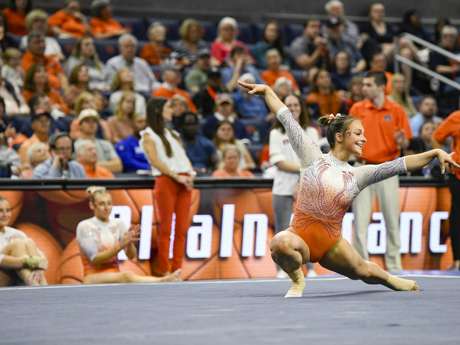 A gymnast in an orange and silver outfit performs a pose while two others sit nearby, with a crowd in the background.