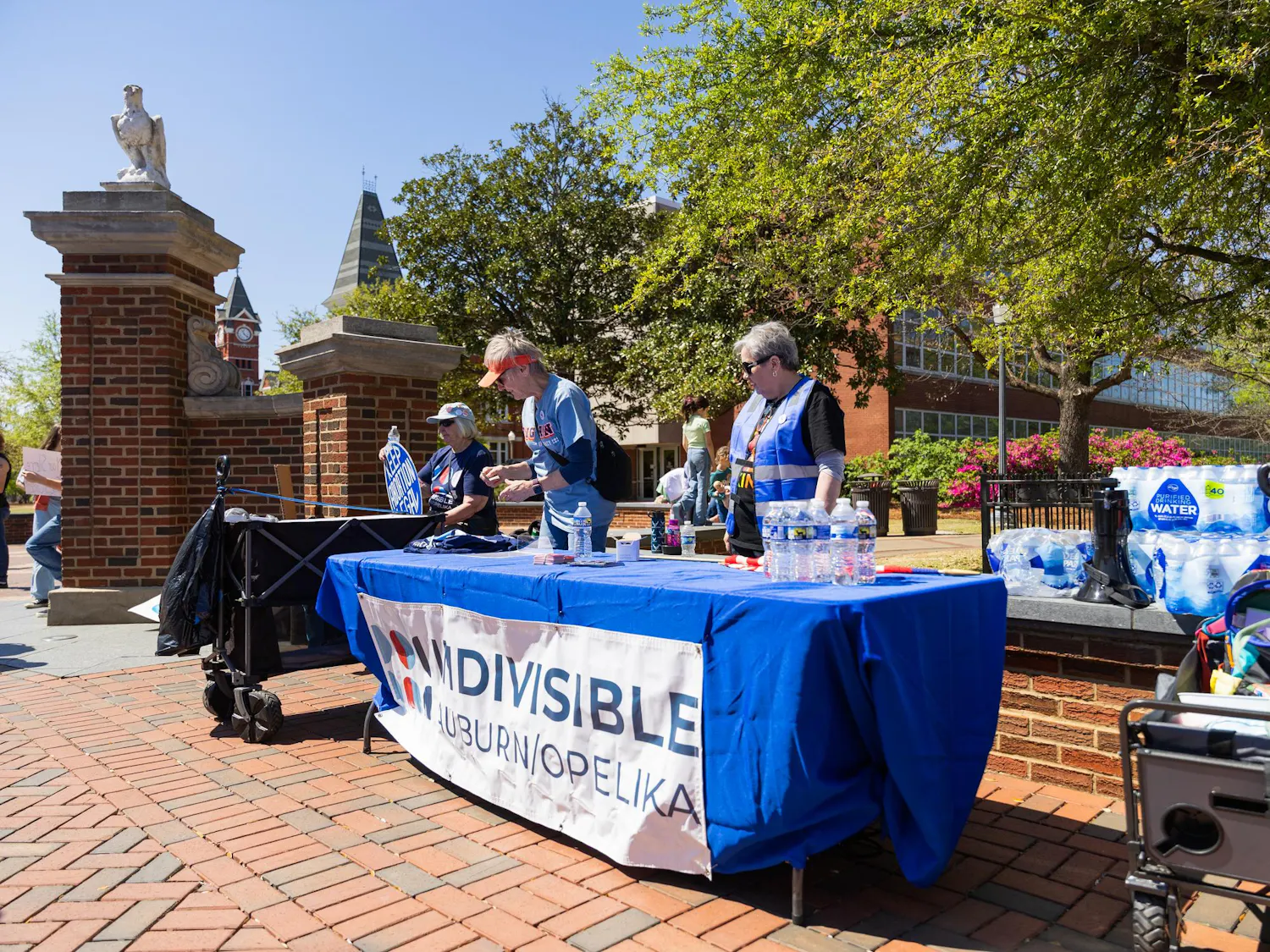A blue table with water bottles and a sign is set up outdoors, while people engage in conversation near brick pathways and trees.