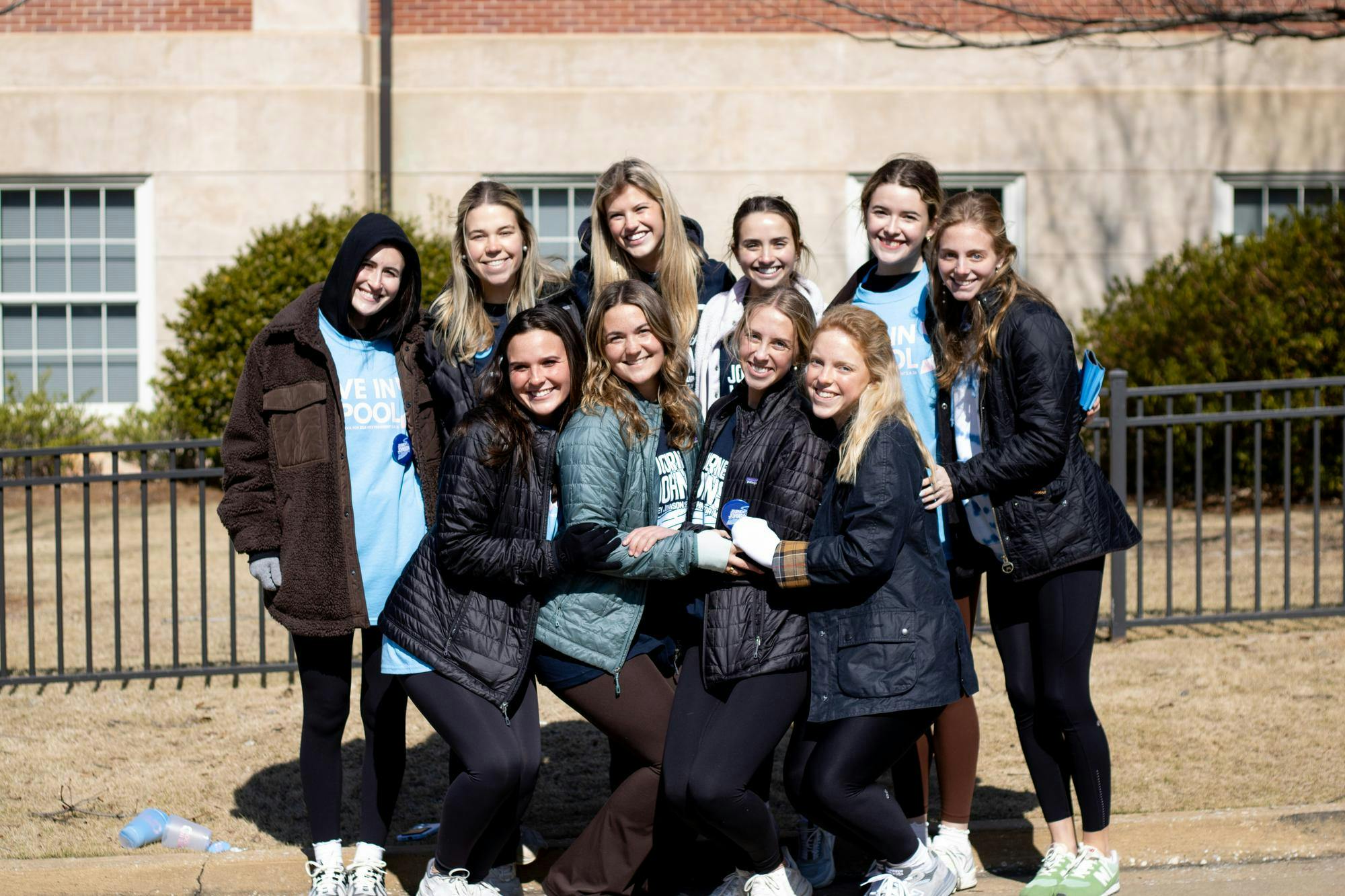 A group of ten young women smiles together outdoors, wearing jackets and fitting leggings, in front of a brick building.