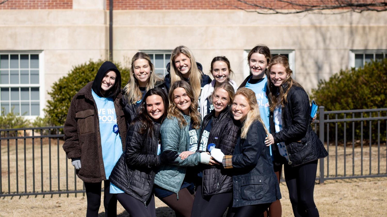 A group of ten young women smiles together outdoors, wearing jackets and fitting leggings, in front of a brick building.