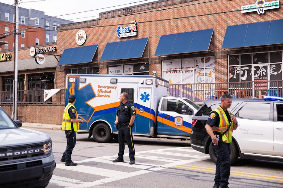 An emergency medical service vehicle with flashing lights is parked near a restaurant, while police officers manage traffic.
