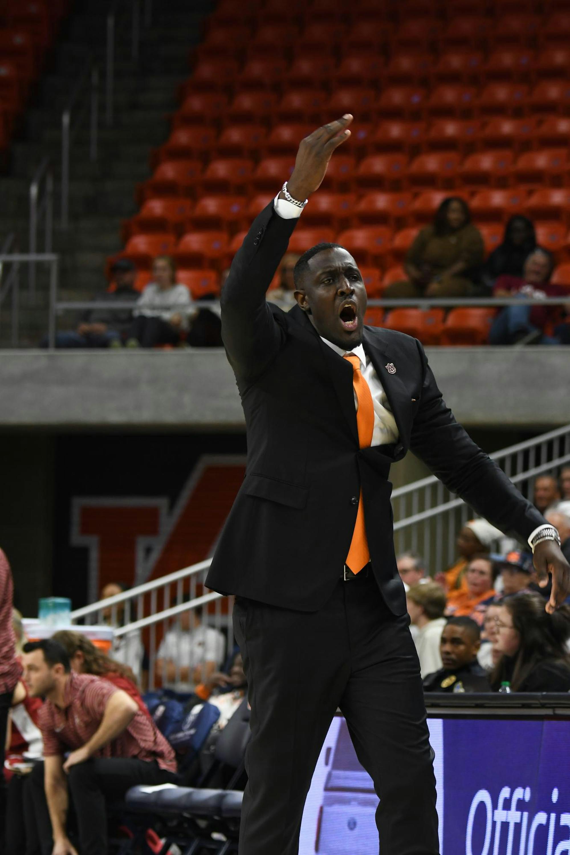 A coach in a suit passionately gestures with one hand raised, surrounded by seated spectators in an arena.