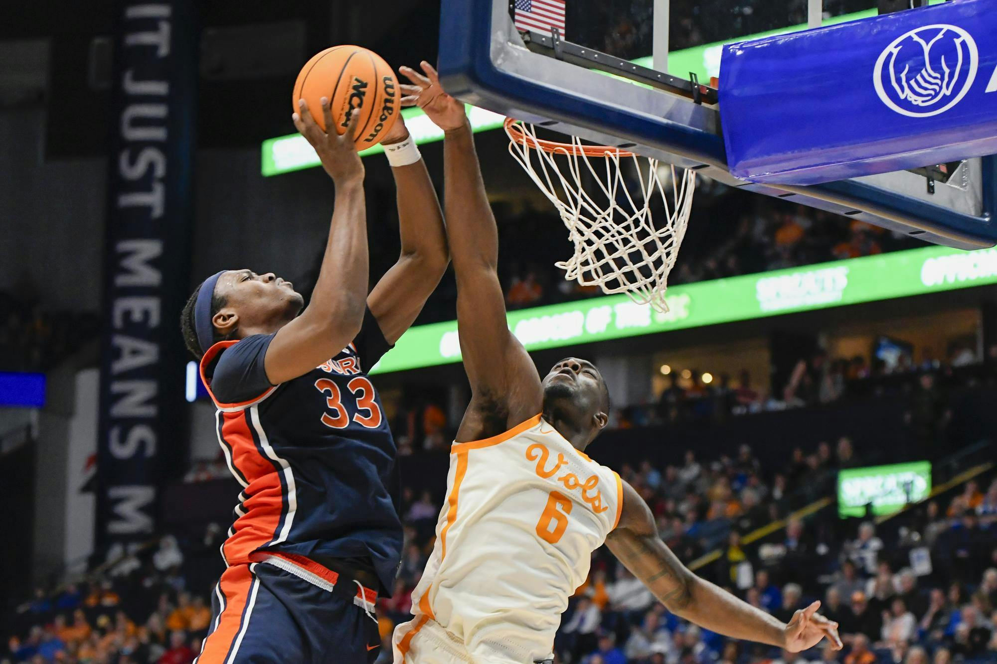 A player in dark uniform jumps to score while another in light uniform attempts to block the shot near the basketball hoop.