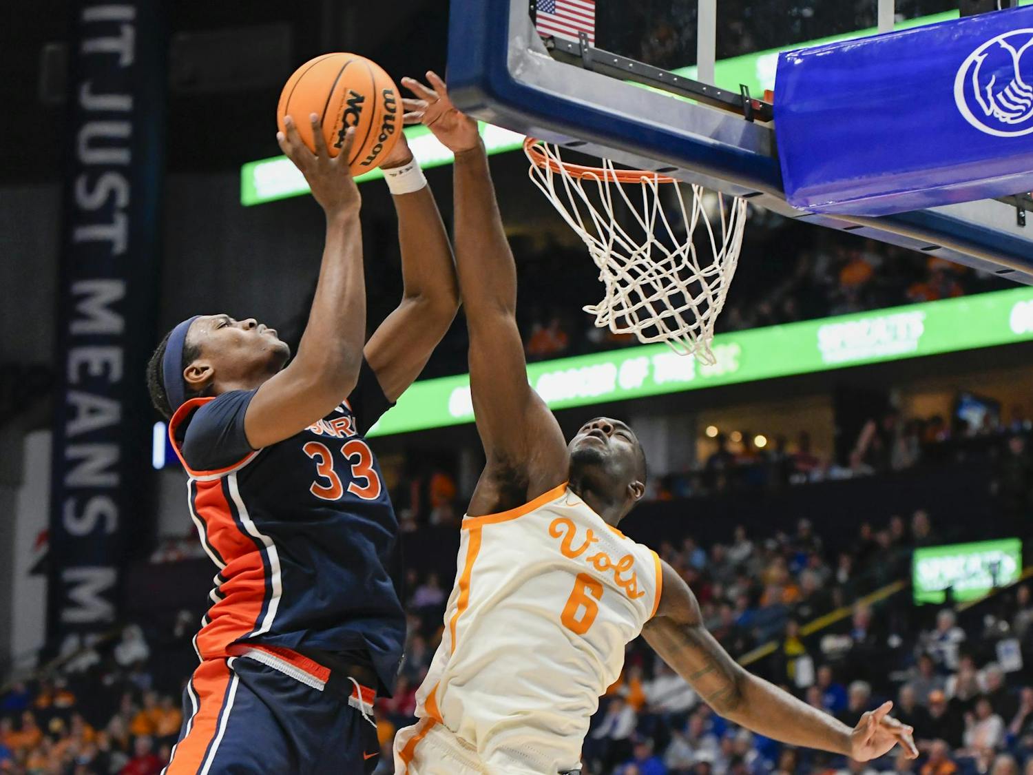 A player in dark uniform jumps to score while another in light uniform attempts to block the shot near the basketball hoop.