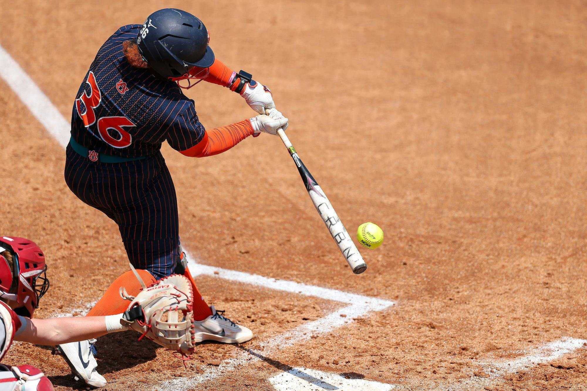 A player swings a bat at a yellow softball while a catcher kneels behind home plate.