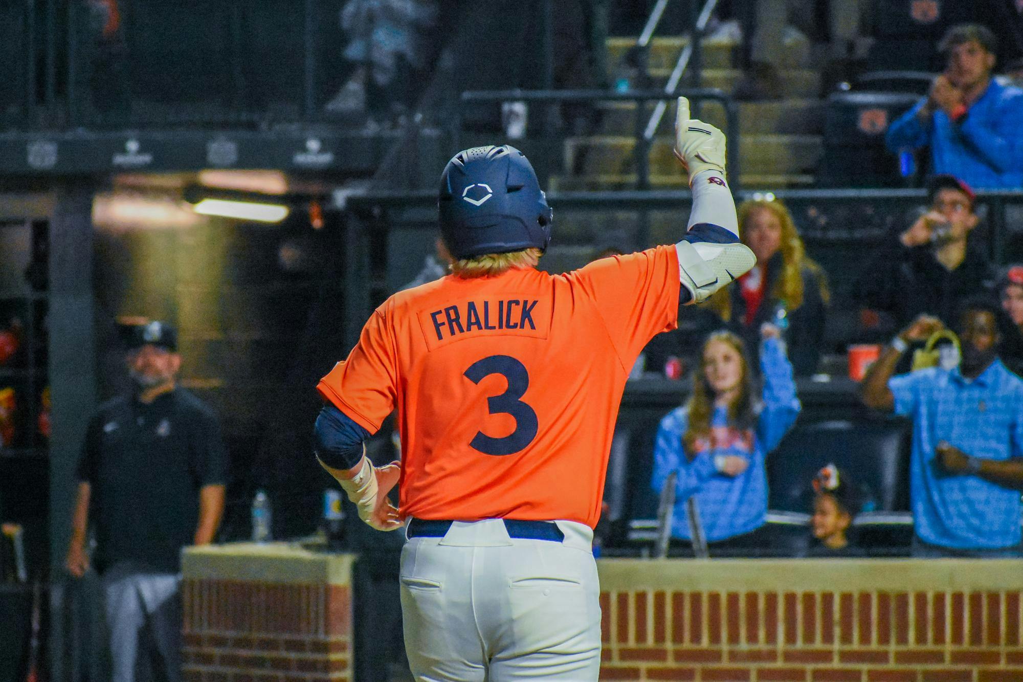 A baseball player in an orange jersey raises one hand while fans watch from the stands.