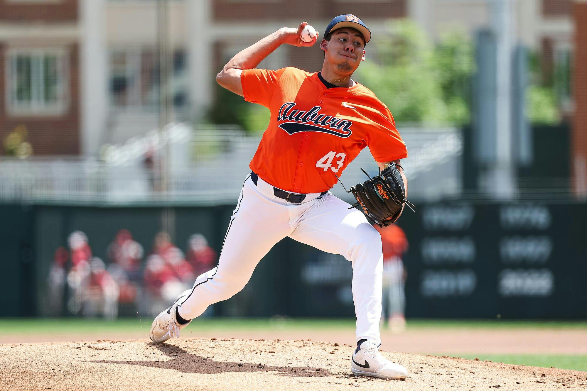 A baseball player in an orange jersey is in mid-pitch on a sunny field, wearing white pants and a black glove.