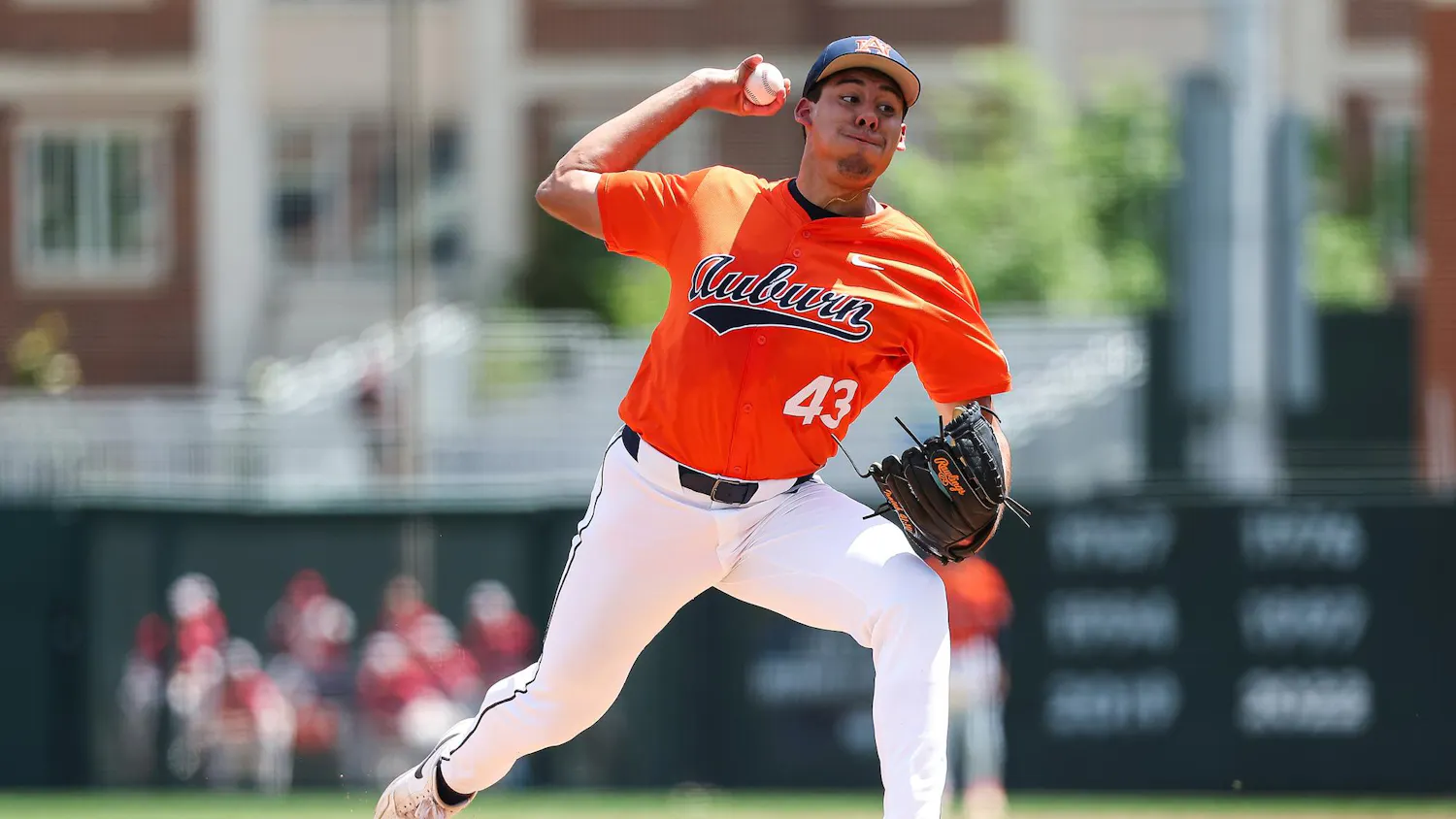 A baseball player in an orange jersey is in mid-pitch on a sunny field, wearing white pants and a black glove.