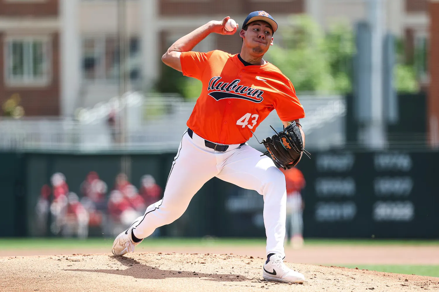 A baseball player in an orange jersey is in mid-pitch on a sunny field, wearing white pants and a black glove.