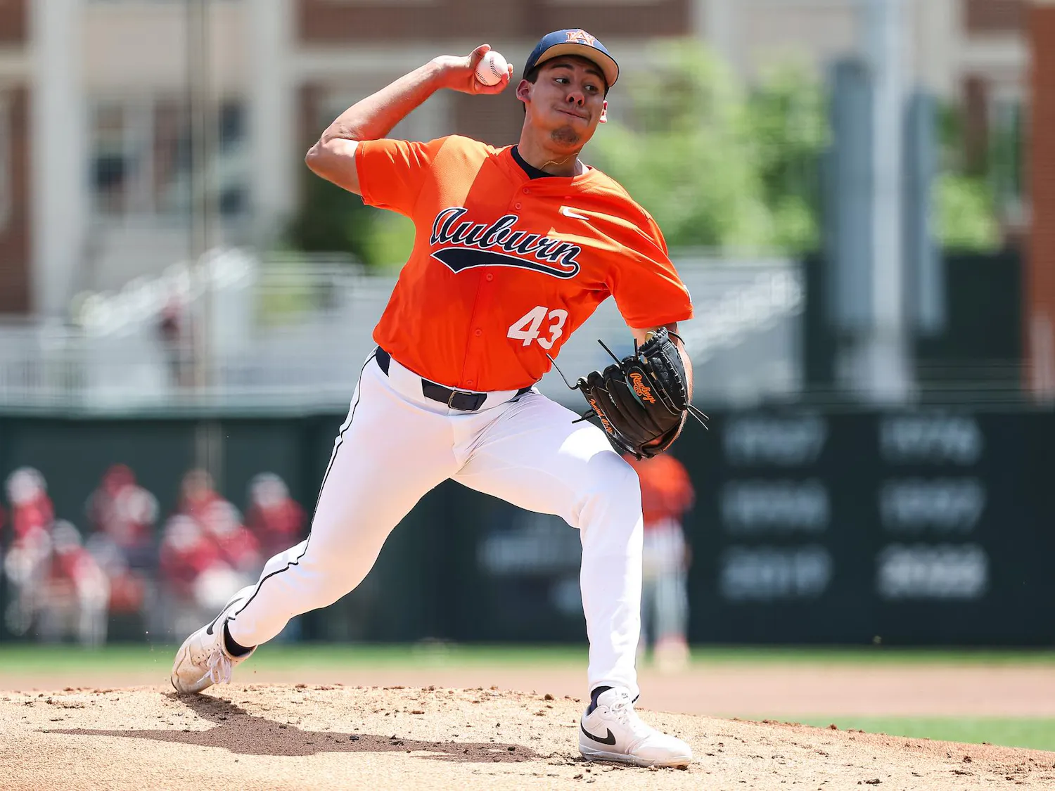 A baseball player in an orange jersey is in mid-pitch on a sunny field, wearing white pants and a black glove.