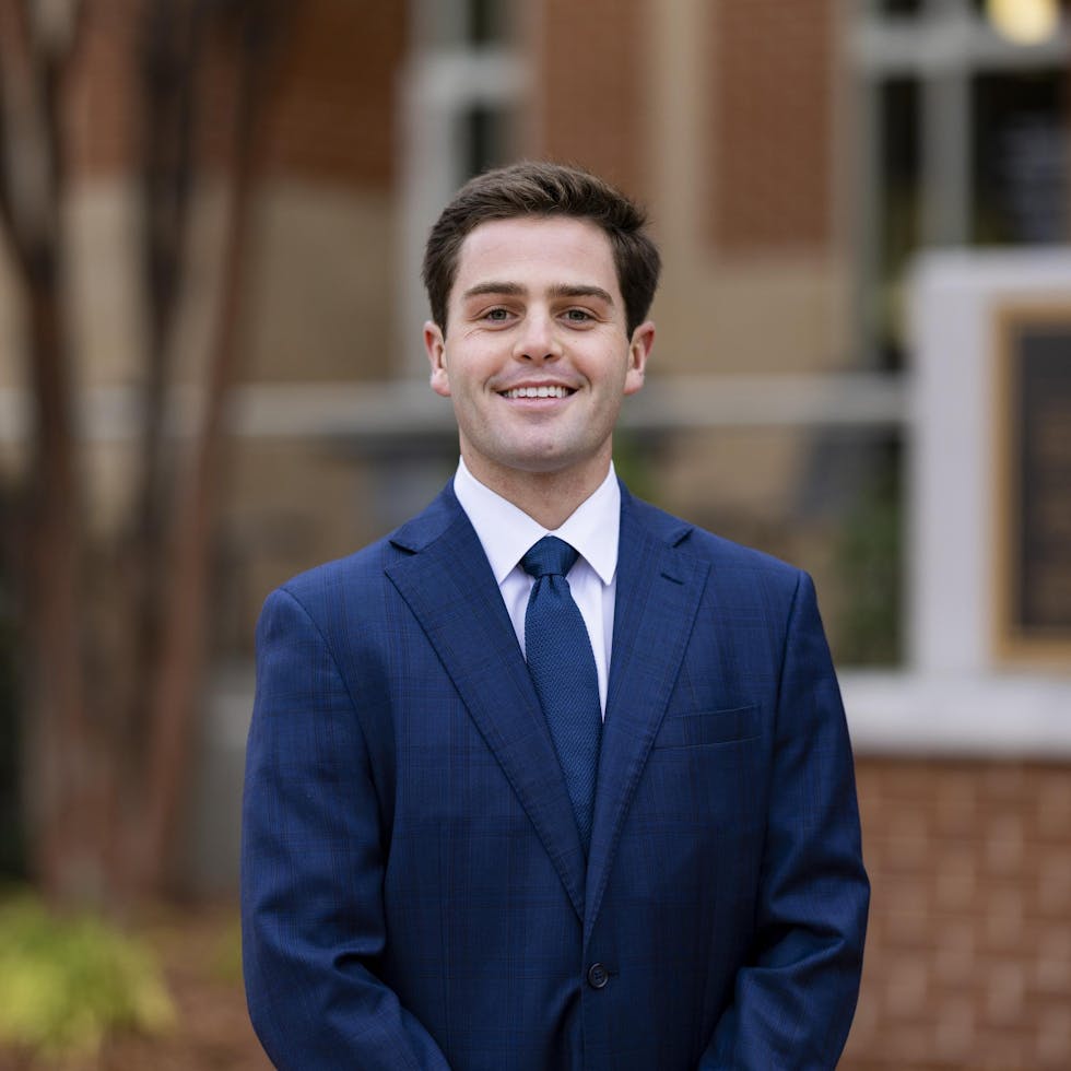 A young man in a blue suit stands confidently outdoors, smiling while looking directly at the camera.