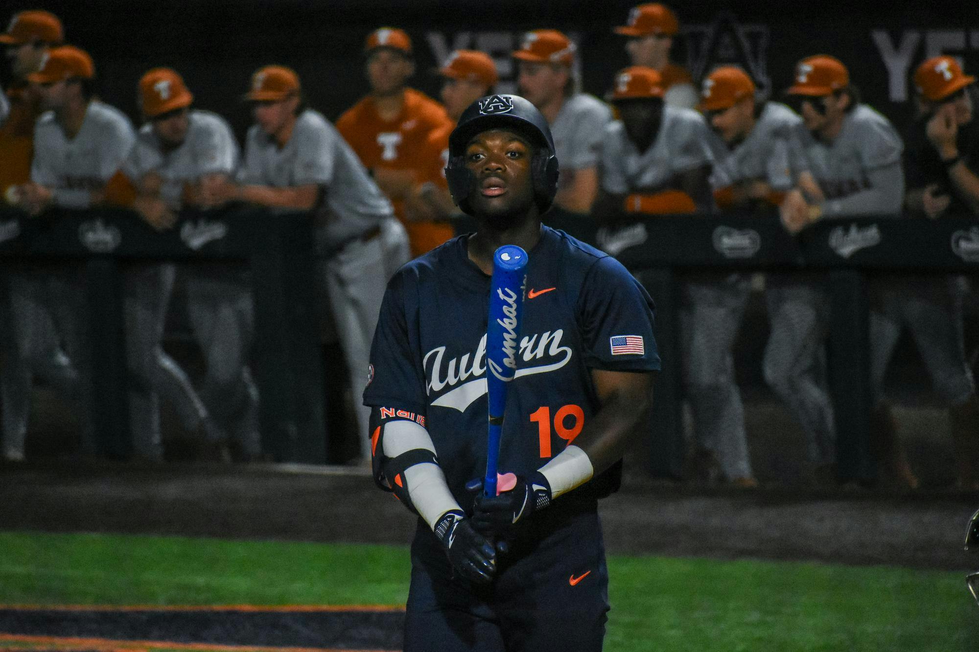A baseball player wearing a dark jersey stands with a bat, while other players in light uniforms observe from the dugout behind him.