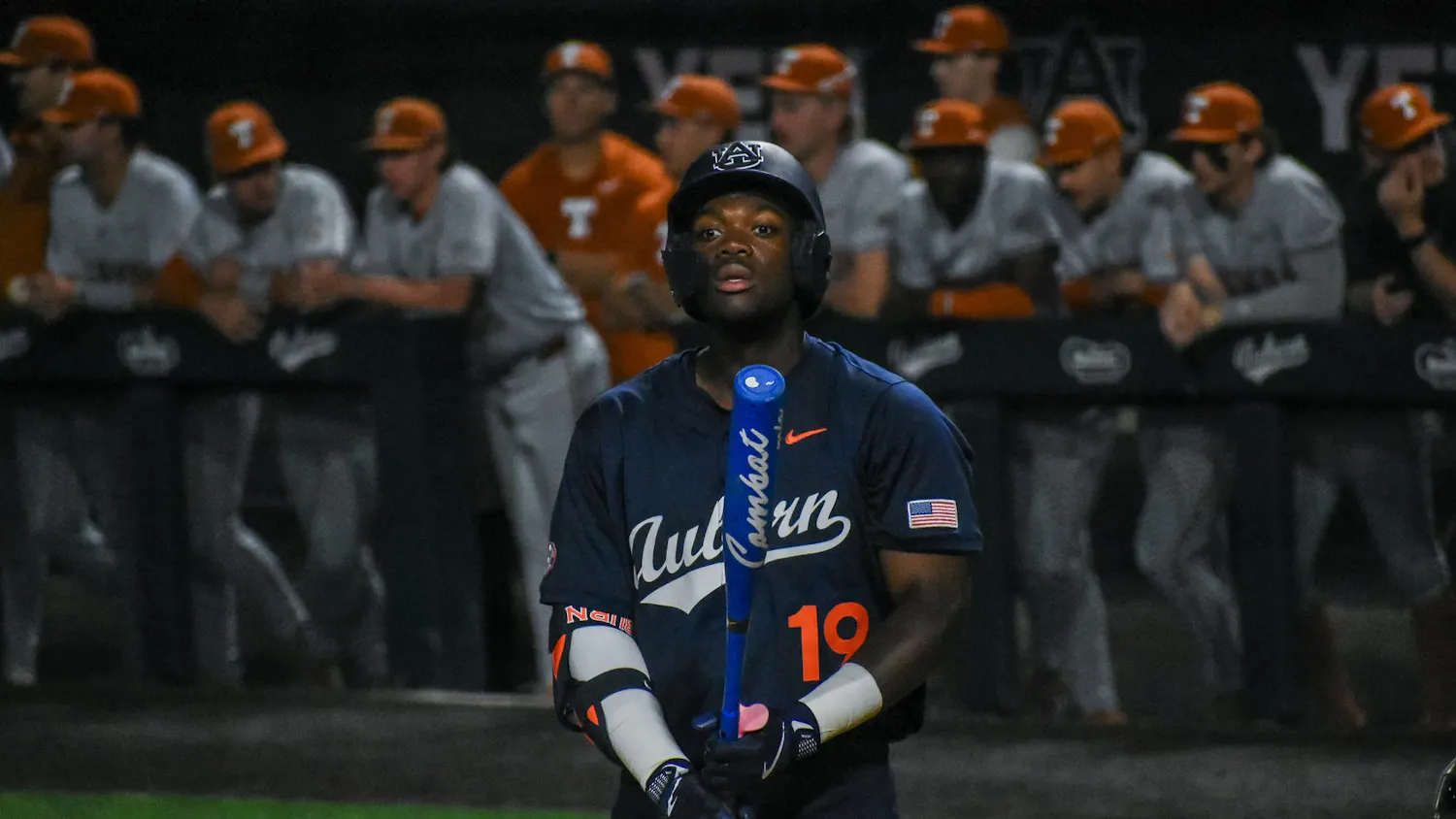A baseball player wearing a dark jersey stands with a bat, while other players in light uniforms observe from the dugout behind him.