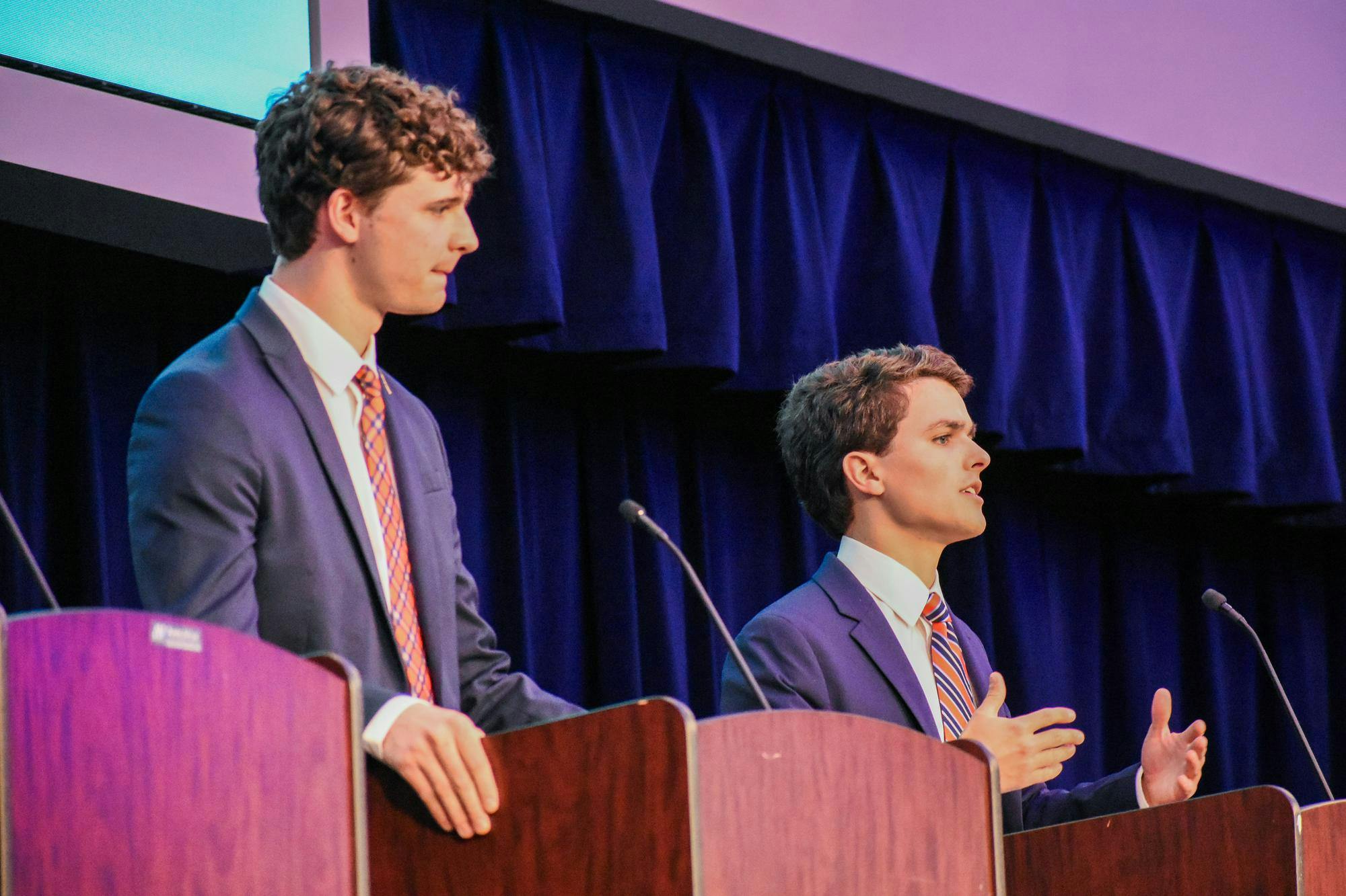 Two young men in suits stand at podiums, speaking into microphones in a formal setting with dark blue curtains behind them.