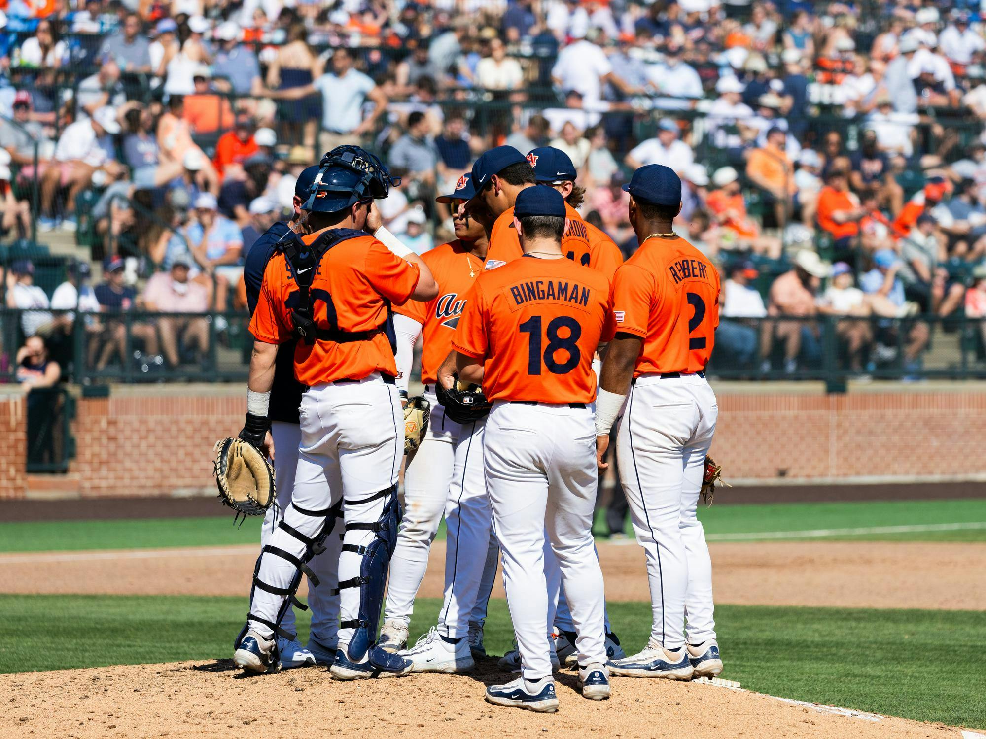 A group of six baseball players in orange jerseys huddles on the pitching mound, surrounded by a large audience in the stands.