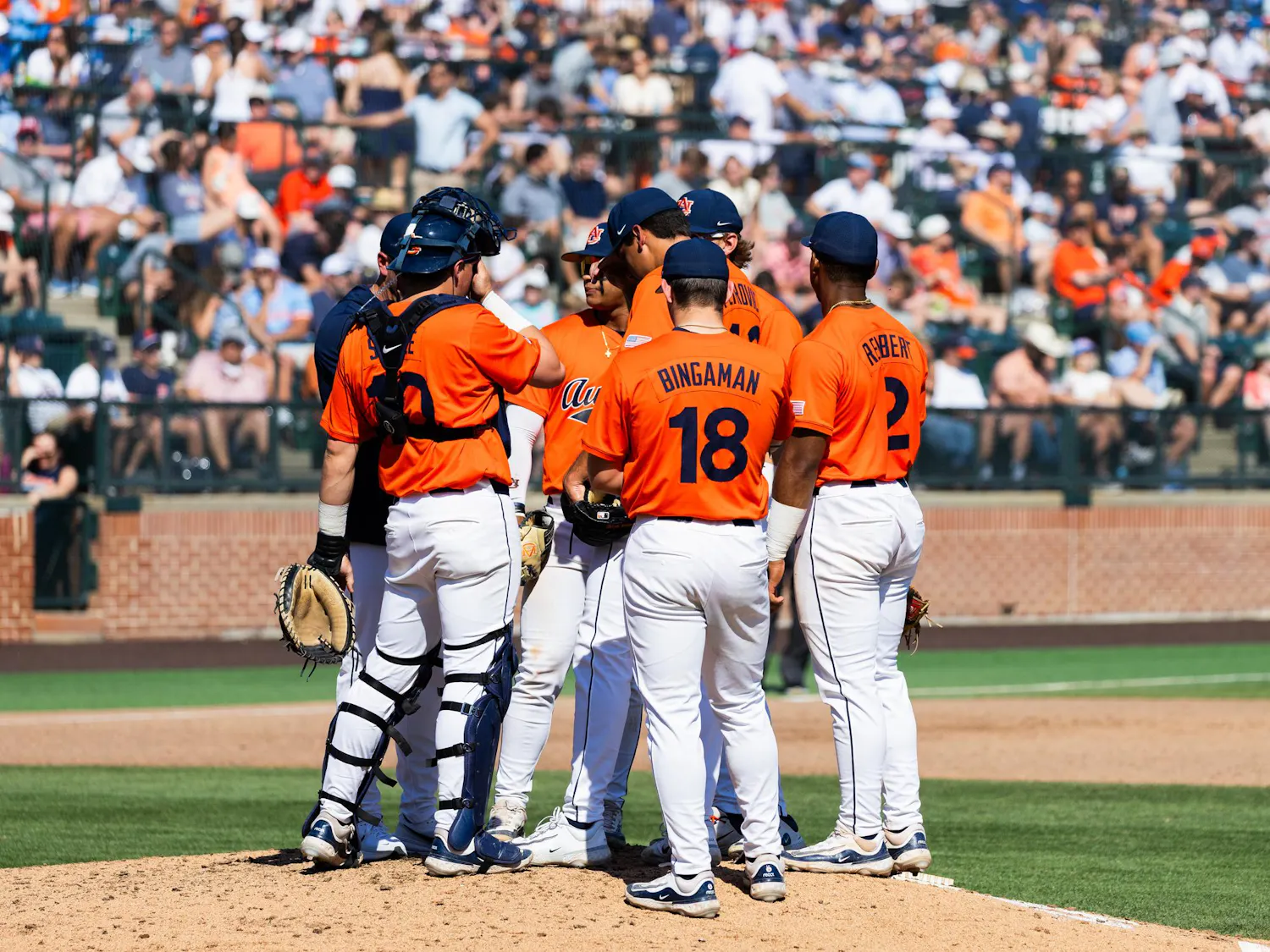A group of six baseball players in orange jerseys huddles on the pitching mound, surrounded by a large audience in the stands.