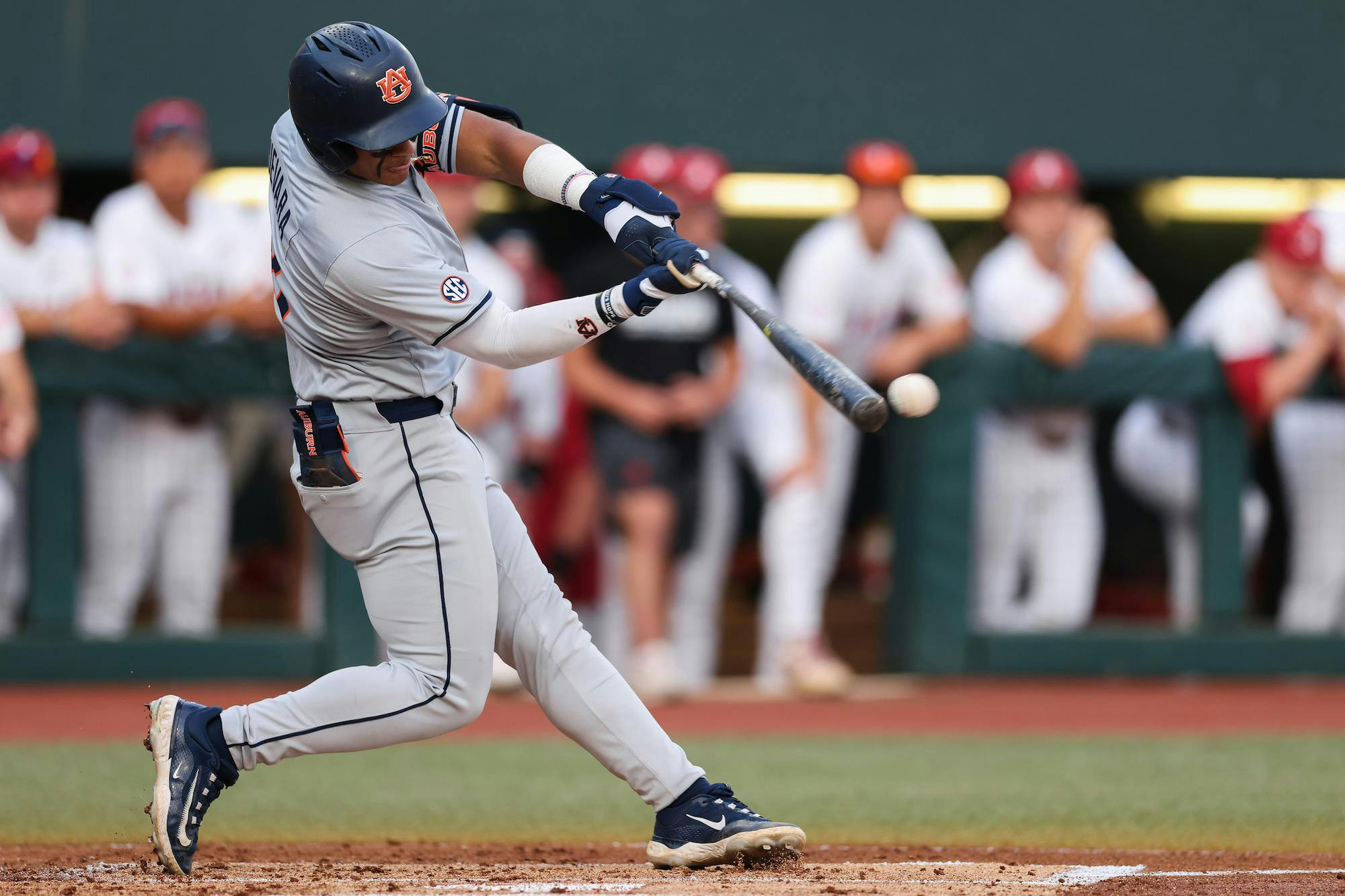 A player in a grey baseball uniform swings a bat at a white baseball, with spectators in the background.