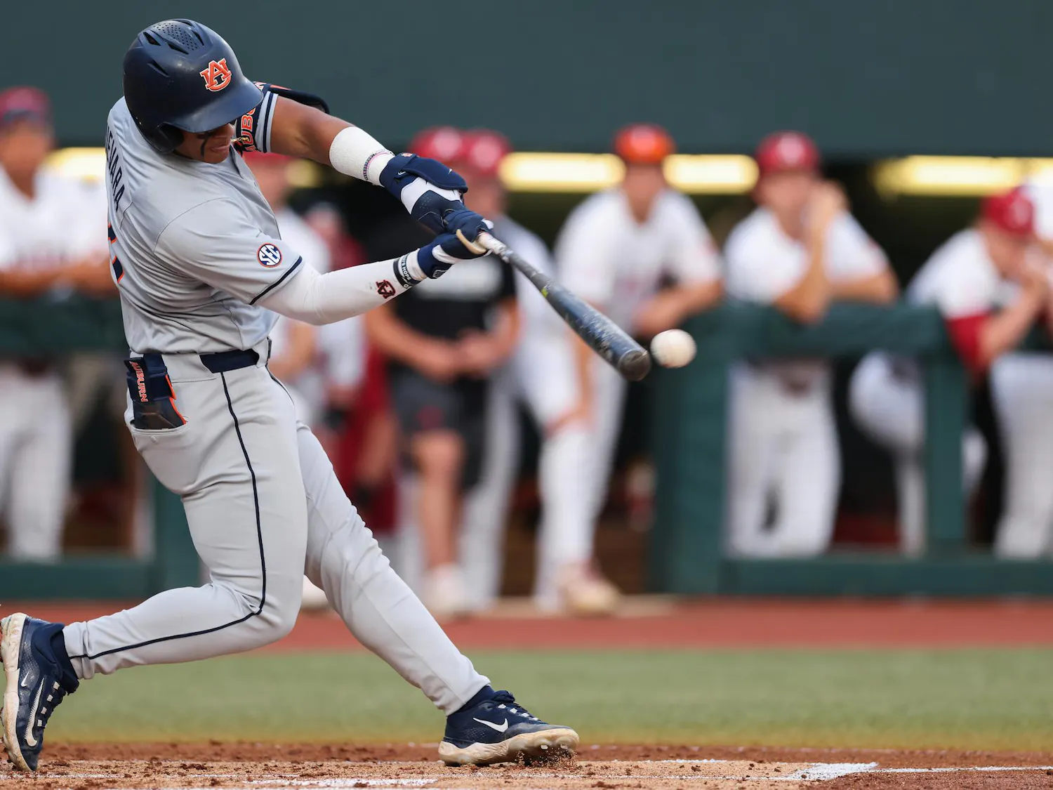 A player in a grey baseball uniform swings a bat at a white baseball, with spectators in the background.