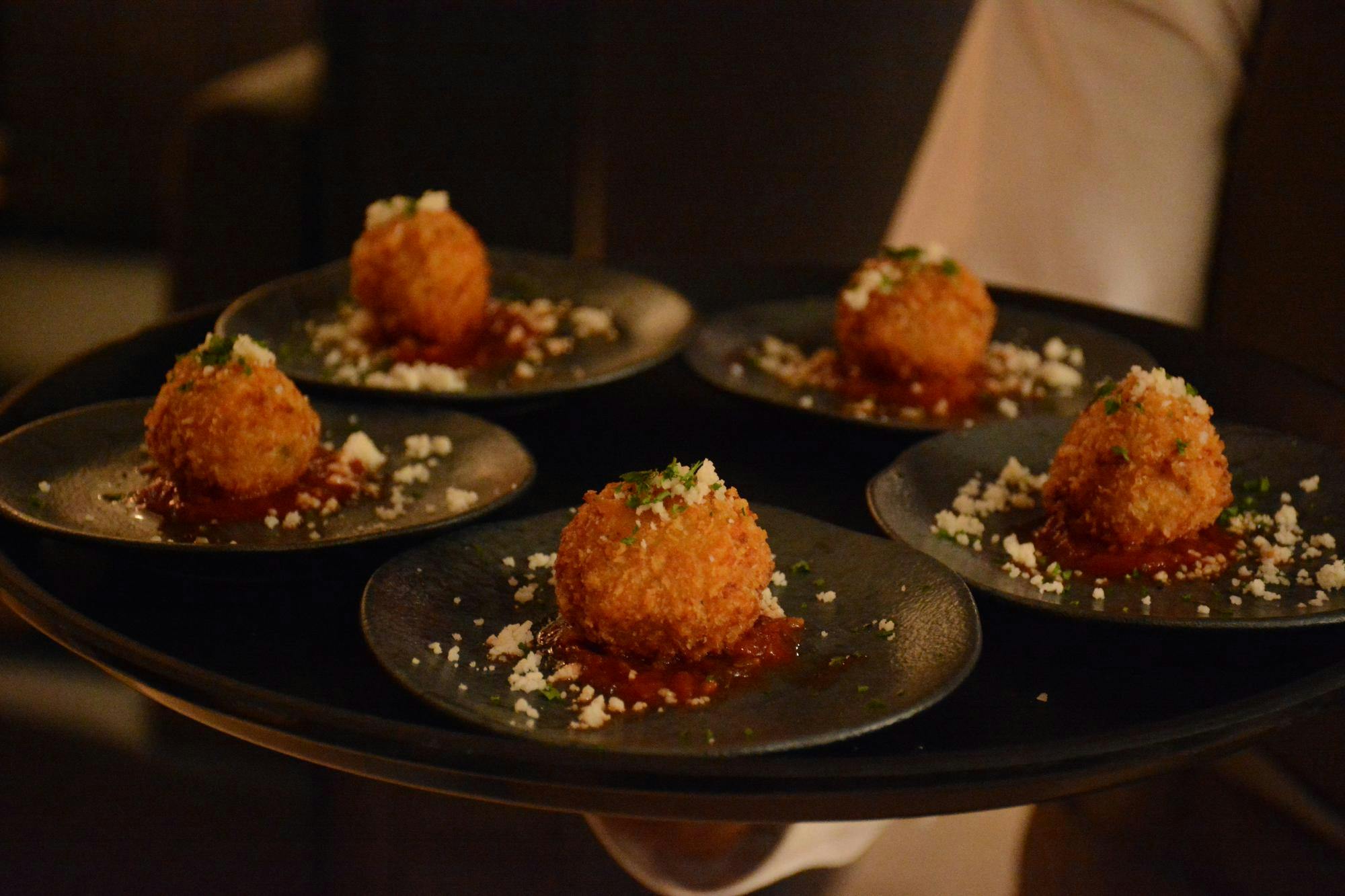 A waiter holds a black tray with five small, golden, crispy balls on plates, topped with white crumbles and garnished with green herbs.