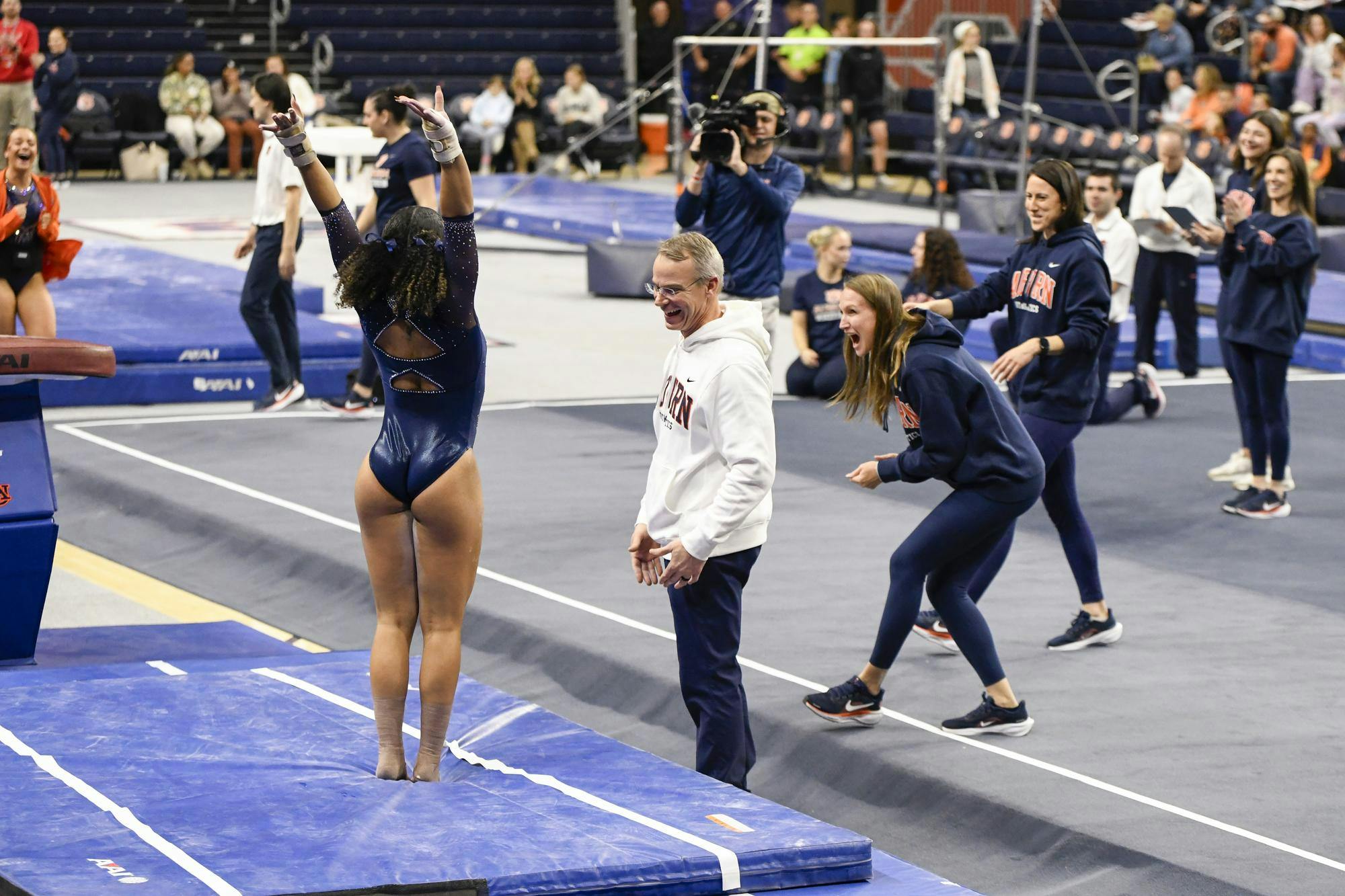 A gymnast celebrates a successful routine while teammates and coaches cheer excitedly in a sports arena.