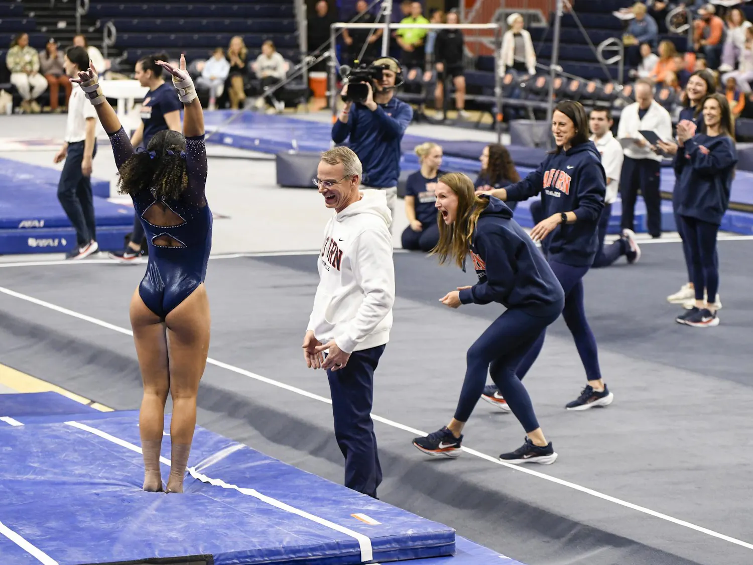 A gymnast celebrates a successful routine while teammates and coaches cheer excitedly in a sports arena.