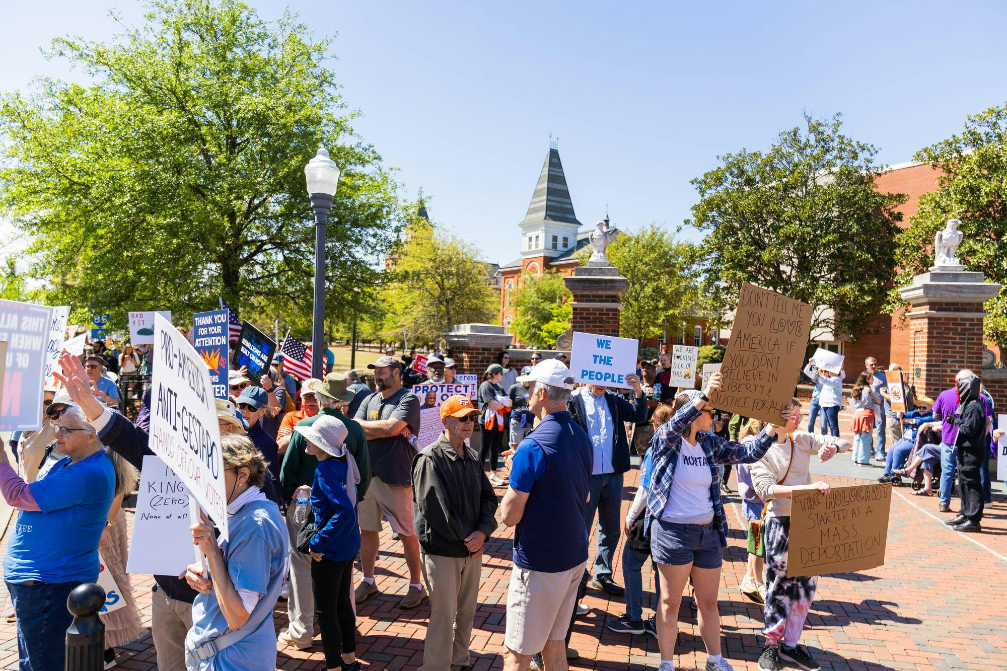 A crowd of protesters holds various signs in a sunny park, with buildings and trees in the background.