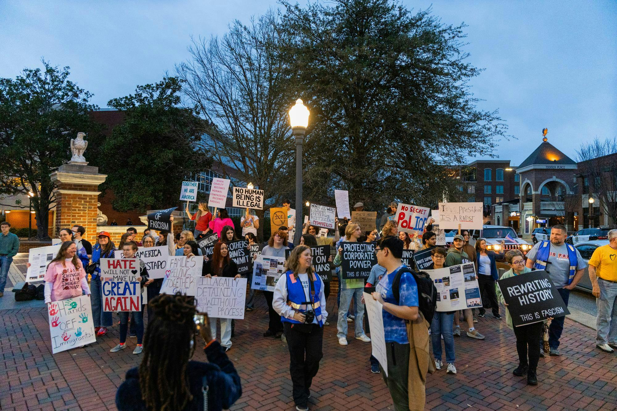 A diverse group of protesters holds signs with various political messages in an outdoor setting during dusk.