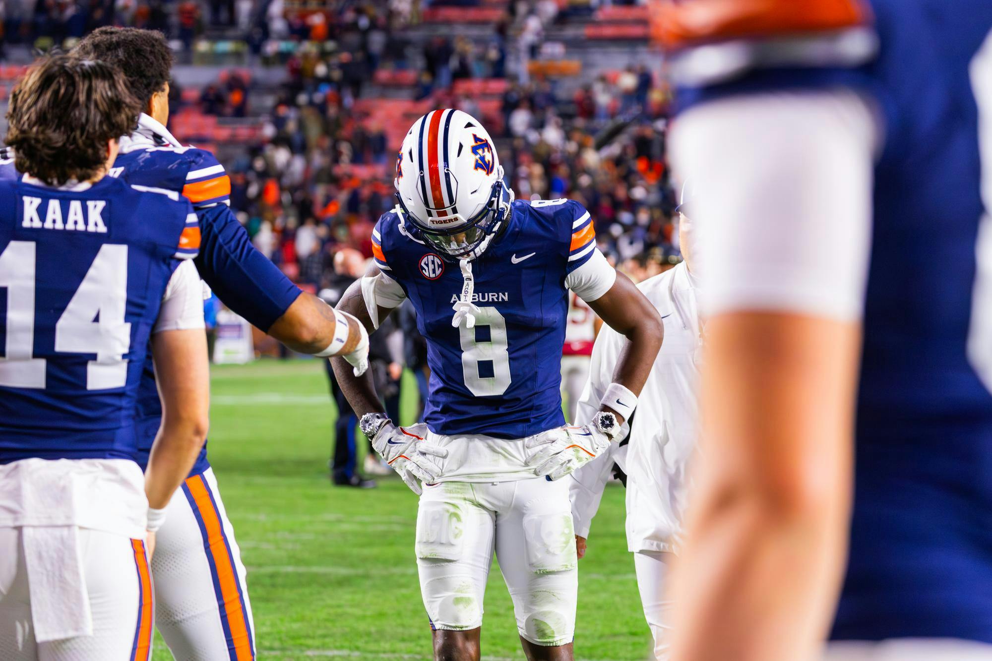 A football player wearing a blue jersey with orange accents bows his head while other players stand nearby, appearing contemplative.