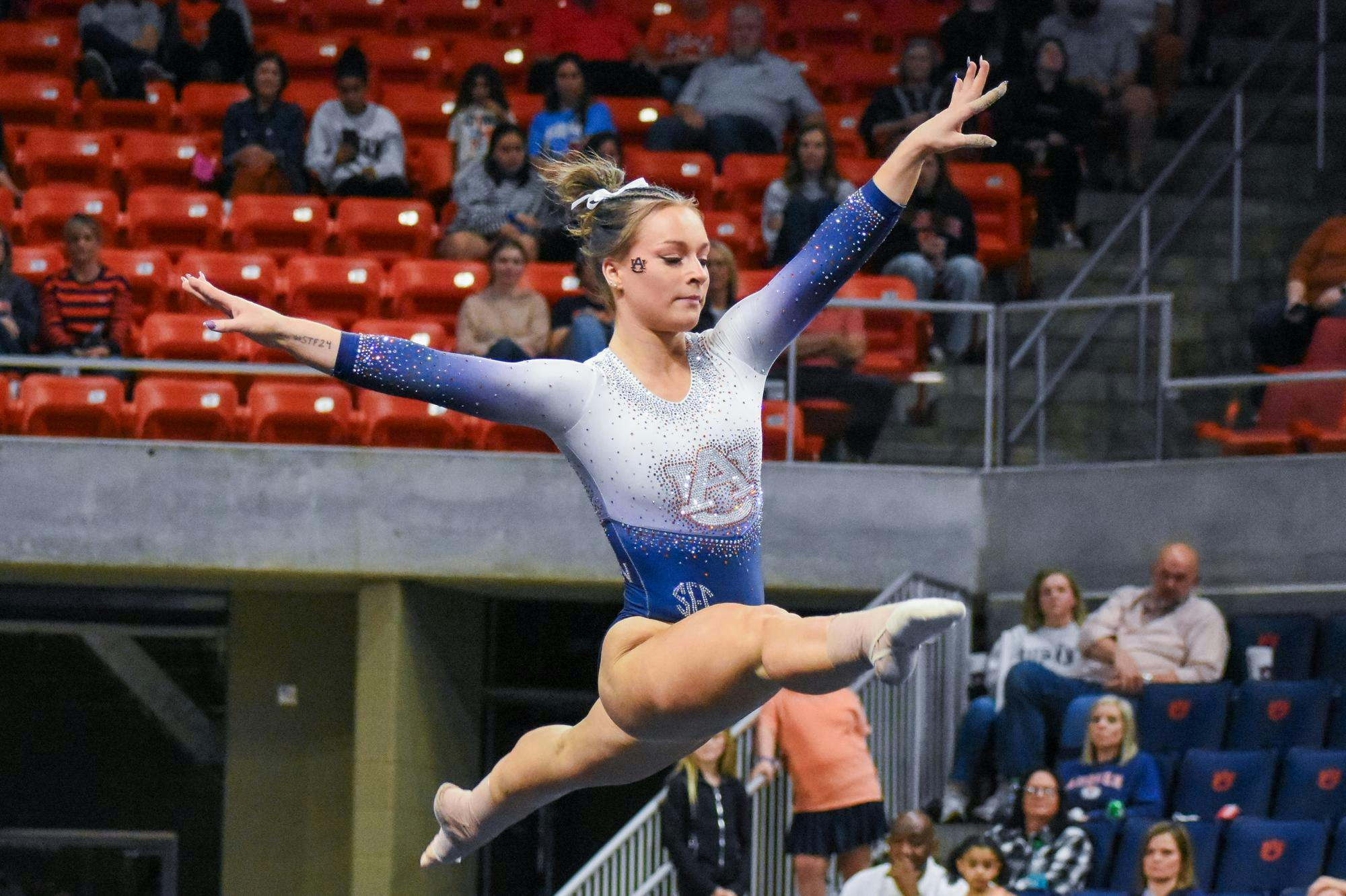 A gymnast in a sparkling blue and white leotard performs a mid-air leap while spectators watch from a stadium.
