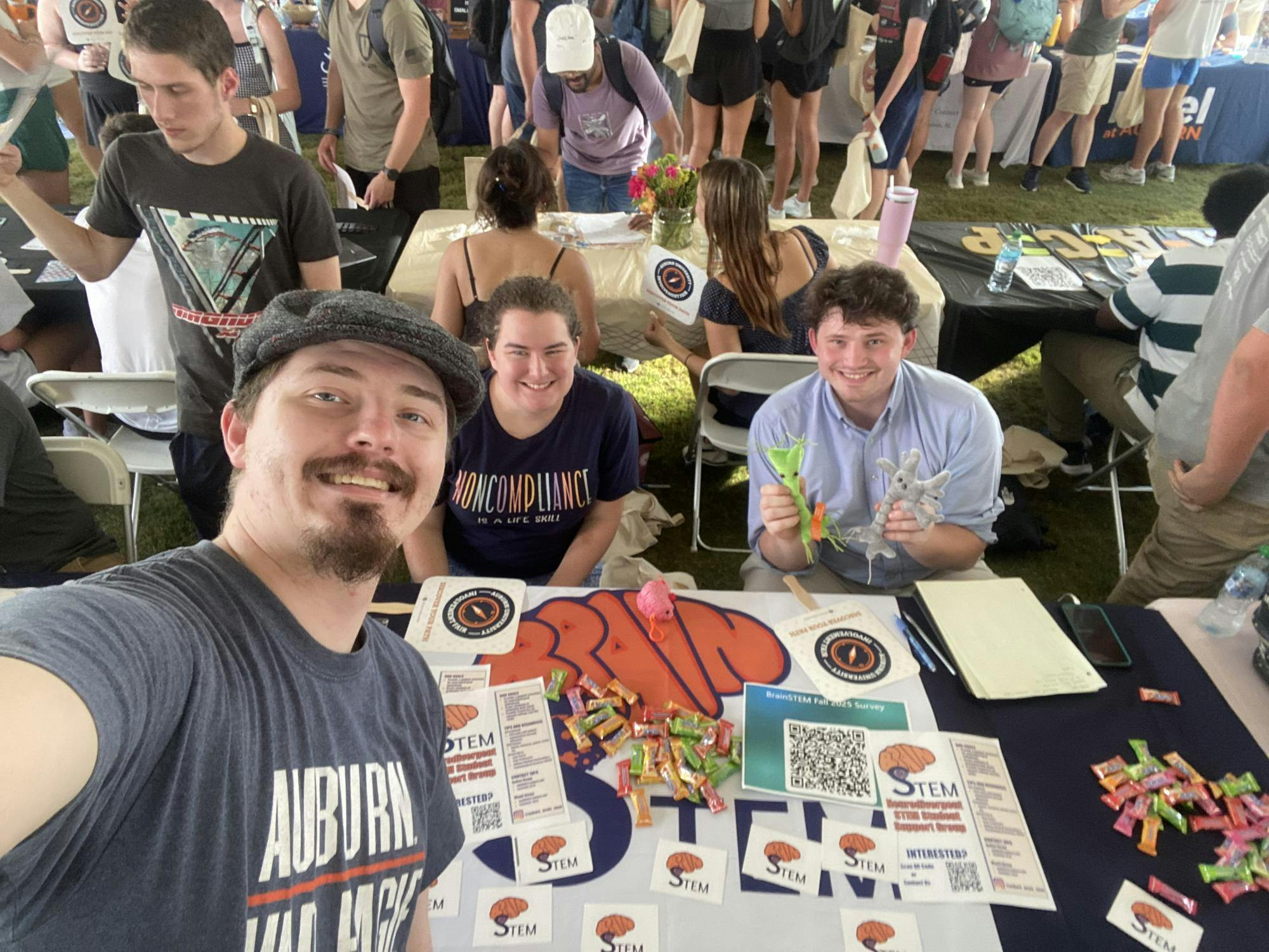A group of people poses at a table filled with colorful snacks, papers and small toys in a crowded outdoor setting.