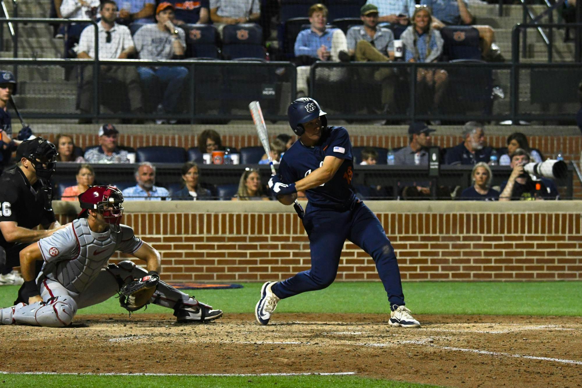 A baseball player in a blue uniform swings a bat while a catcher in gray crouches behind home plate, with spectators in the stands.