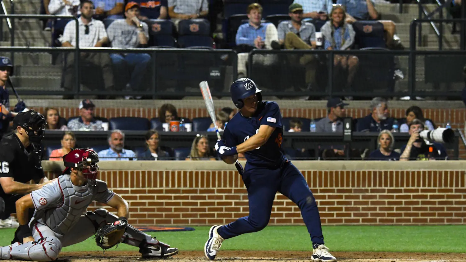 A baseball player in a blue uniform swings a bat while a catcher in gray crouches behind home plate, with spectators in the stands.