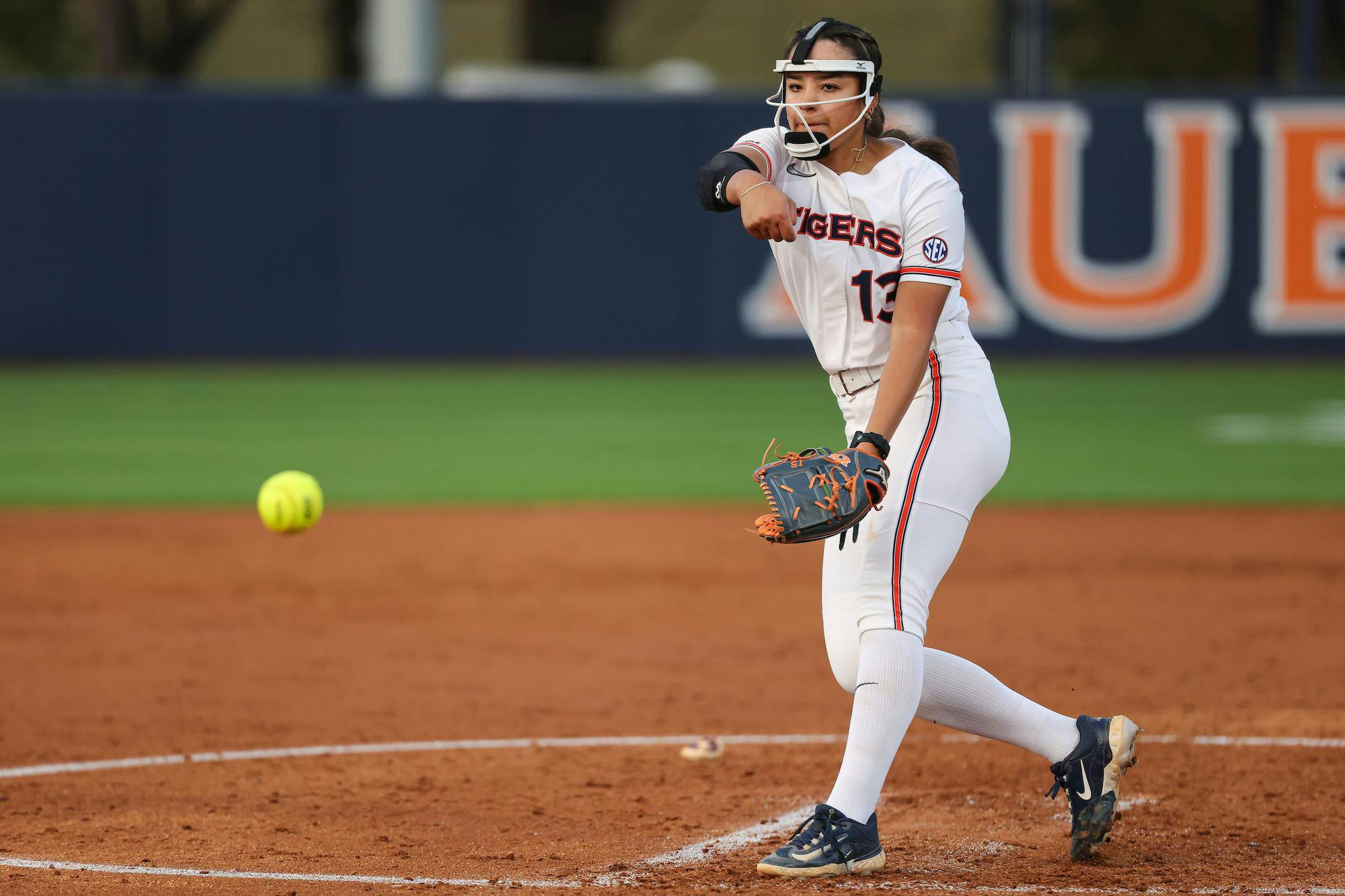 A female softball pitcher in a white uniform throws a yellow ball toward home plate on a dirt field.
