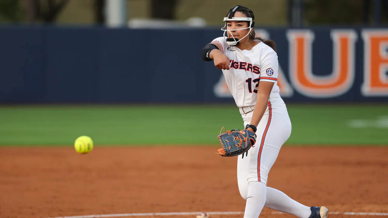 A female softball pitcher in a white uniform throws a yellow ball toward home plate on a dirt field.