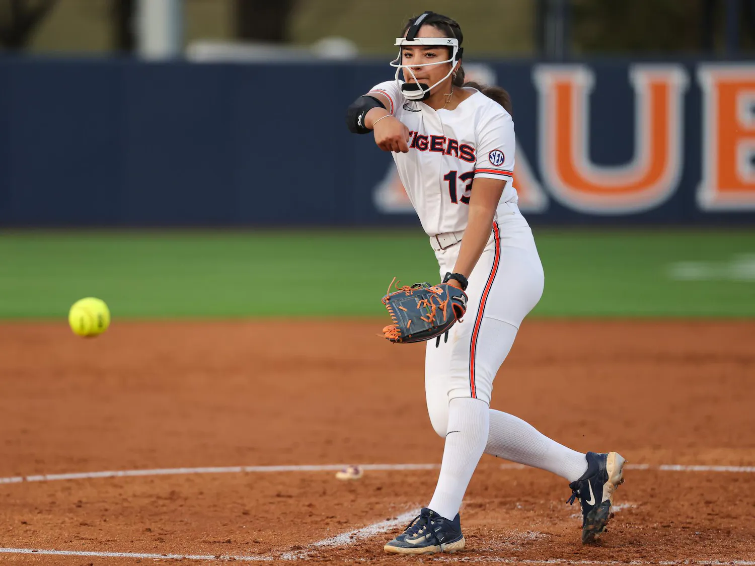 A female softball pitcher in a white uniform throws a yellow ball toward home plate on a dirt field.