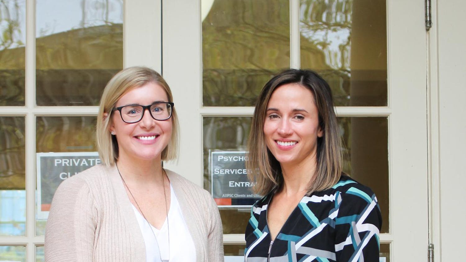 Two women stand together in front of glass doors with signs, one wearing a light sweater and the other a patterned blouse.