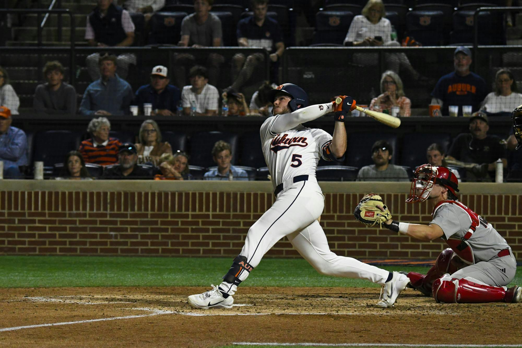 A baseball player swings a bat while a catcher squats behind home plate, with spectators in the stands watching intently.