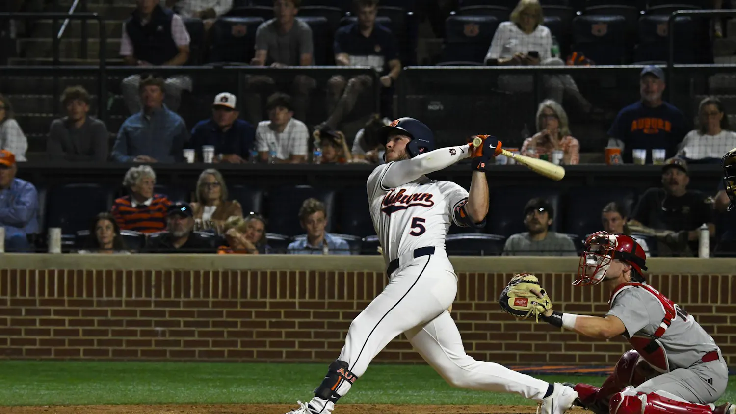 A baseball player swings a bat while a catcher squats behind home plate, with spectators in the stands watching intently.