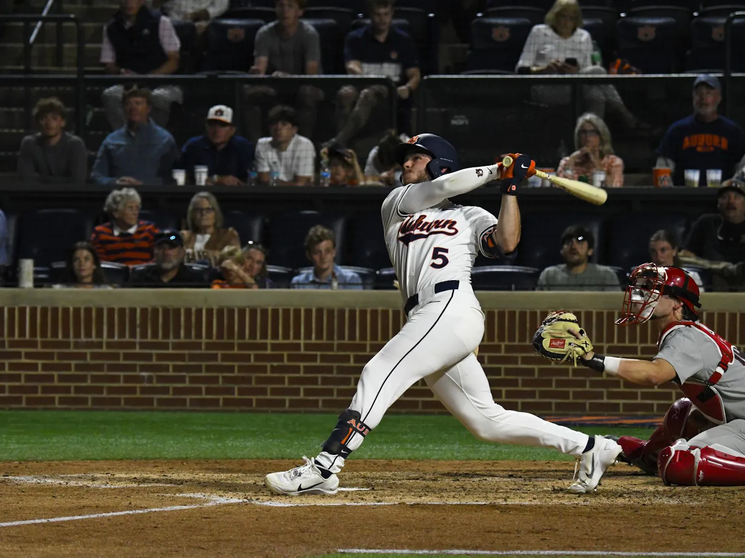 A baseball player swings a bat while a catcher squats behind home plate, with spectators in the stands watching intently.