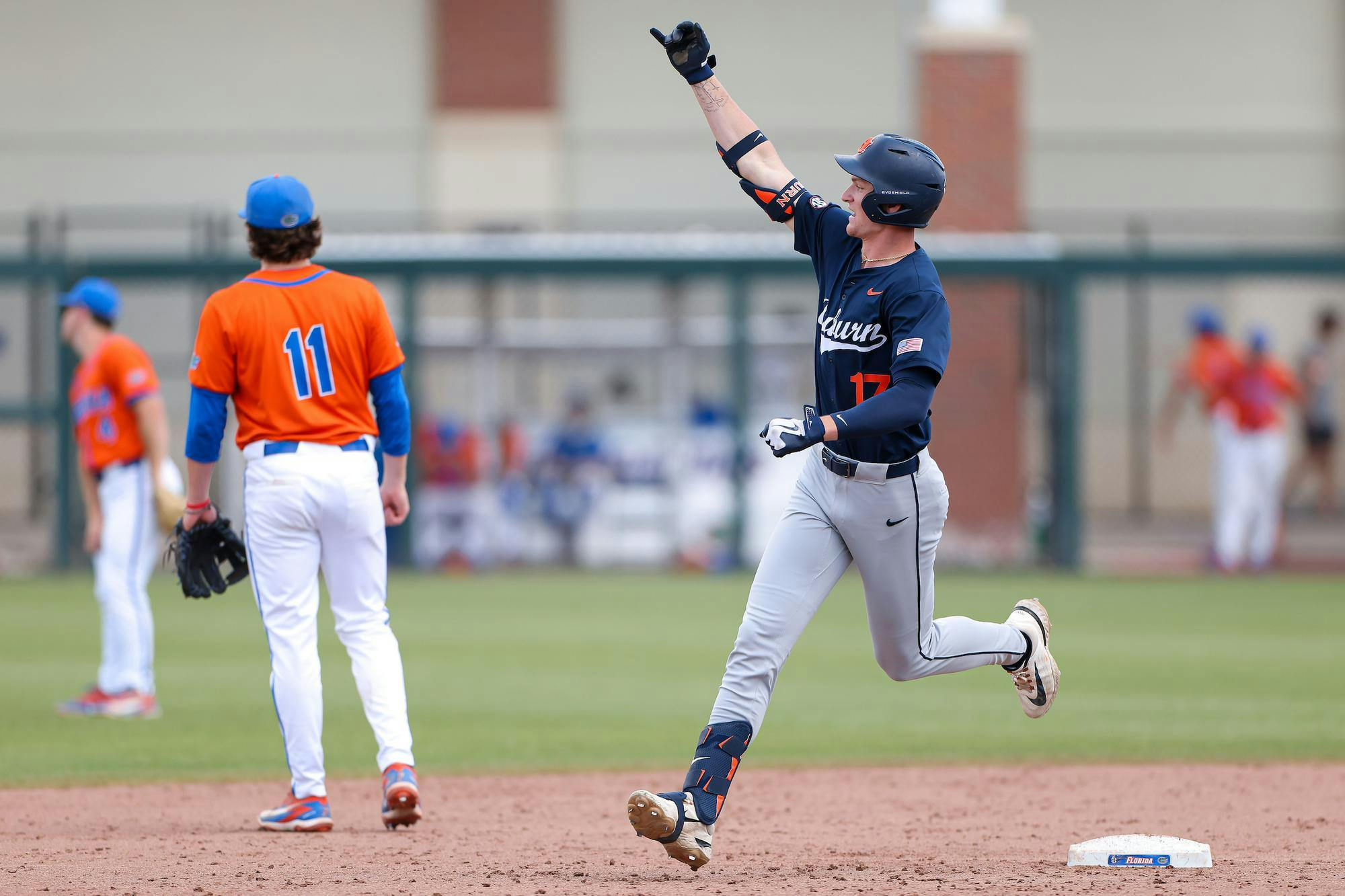 A player in a navy uniform celebrates while running towards a base, with teammates in orange uniforms watching nearby.