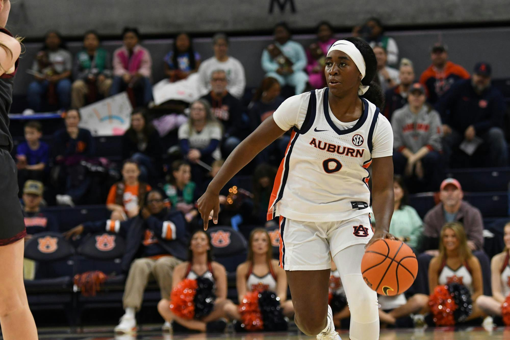 A player in a white basketball uniform dribbles a ball on court, while spectators watch and cheer from the stands.