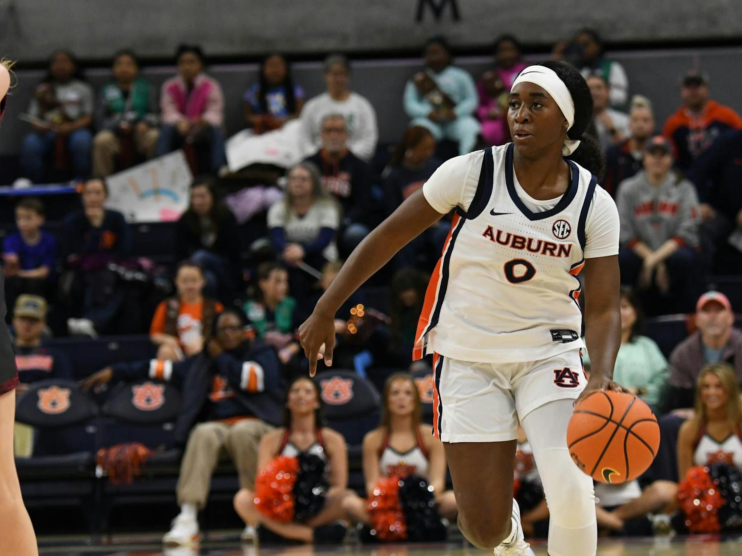 A player in a white basketball uniform dribbles a ball on court, while spectators watch and cheer from the stands.