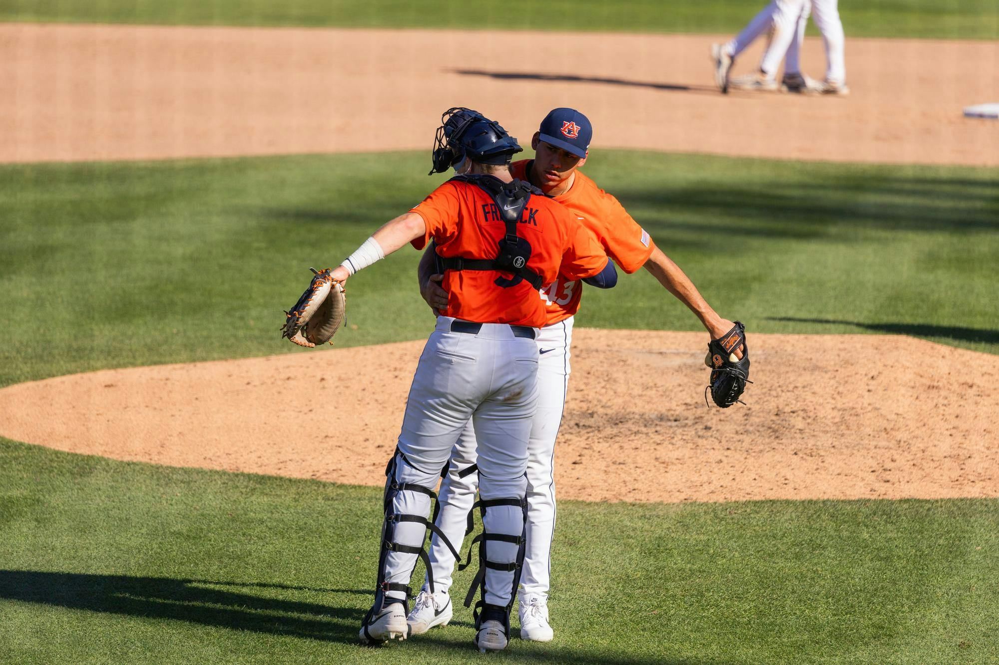 Two baseball players in orange jerseys embrace on the field, with a pitcher in white pants and a catcher wearing gear.